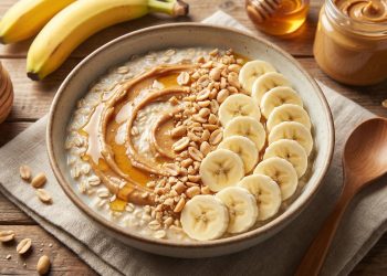 Keramička zdjela s proteinskim bowlom od zobenih pahuljica prelivena spiralom kikiriki maslaca i medom, ukrašena urednim redovima ploškica banane i sjeckanim kikirikijem, poslužena na drvenoj podlozi s lanenom salvetom, uz staklenku kikiriki maslaca i banane u pozadini.