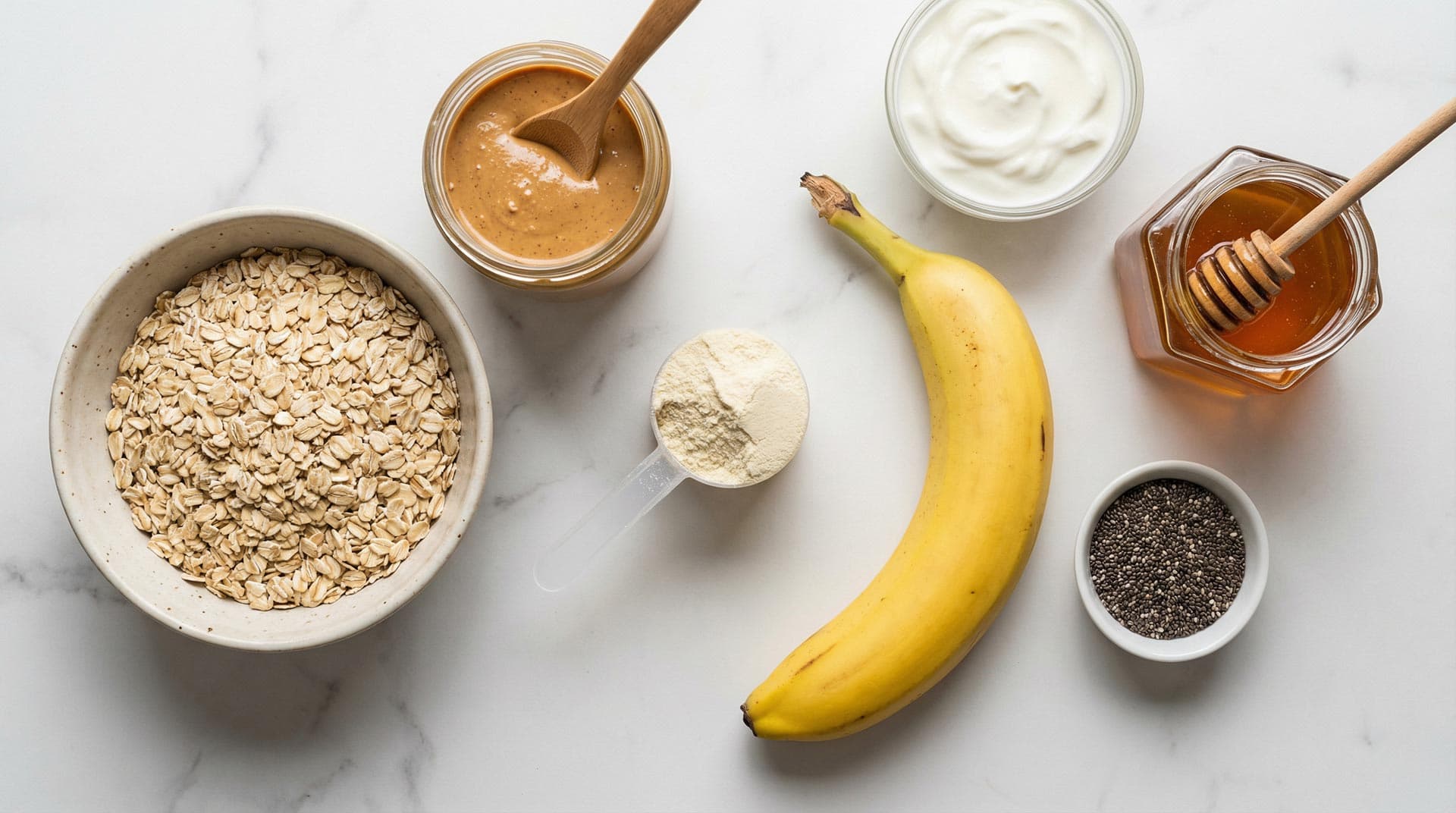 Flat lay prikaz sastojaka za proteinski bowl na bijelom mramoru: zdjela zobenih pahuljica, staklenka kikiriki maslaca s drvenom žlicom, mjerna žlica whey protein praha, cijela banana, zdjela grčkog jogurta, posudica chia sjemenki i staklenka meda s medenim štapićem.